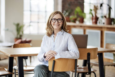 Smiling woman wearing eyeglasses sitting on chair at home - FMKF07579