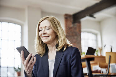 Smiling businesswoman wearing blazer using smart phone at home - FMKF07588