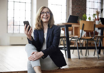 Happy businesswoman with smart phone sitting on hardwood floor - FMKF07590