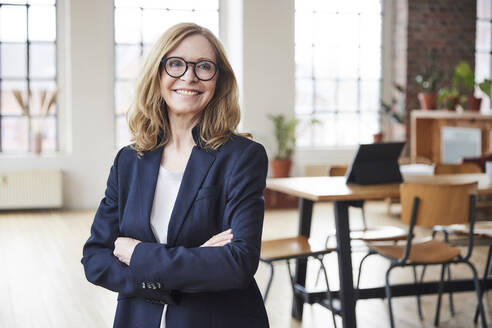 Happy businesswoman wearing eyeglasses standing with arms crossed at home - FMKF07591