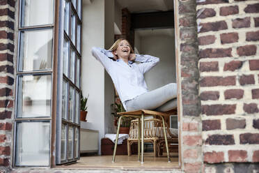 Happy woman sitting on chair at terrace - FMKF07596