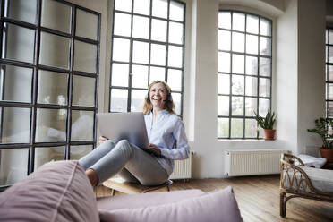 Happy freelancer with headset and laptop sitting in front of window at home - FMKF07600