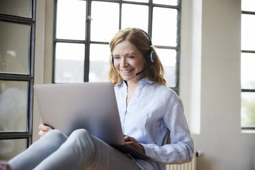 Glücklicher Freiberufler mit Headset bei der Arbeit am Laptop zu Hause - FMKF07601