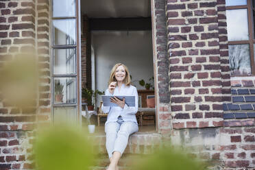 Smiling businesswoman with tablet PC sitting on doorway - FMKF07605