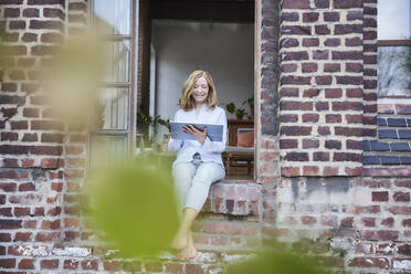 Smiling businesswoman using tablet PC sitting on doorway - FMKF07606
