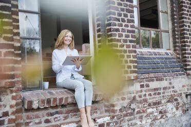 Happy freelancer with tablet PC sitting on doorway - FMKF07607
