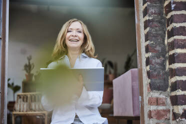 Glückliche Frau mit Tablet-PC im Türrahmen sitzend - FMKF07608