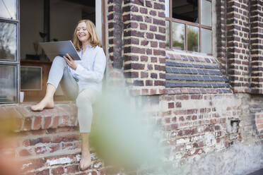 Happy freelancer using digital tablet sitting on doorway - FMKF07611