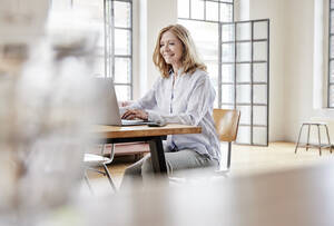 Lächelnder Freiberufler mit Laptop am Tisch im Heimbüro - FMKF07618