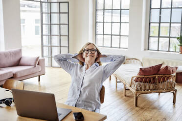 Smiling freelancer with hands behind head sitting on chair at home office - FMKF07622