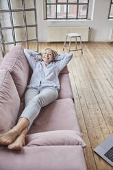 Happy woman with hands behind head lying on sofa at home - FMKF07625