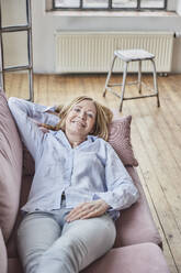 Smiling woman with hand behind head lying on sofa at home - FMKF07626