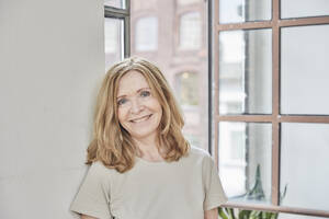 Smiling woman leaning on wall in front of window at home - FMKF07634