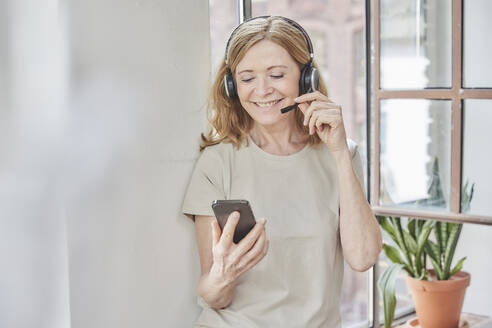 Smiling woman with headset using smart phone standing in front of window at home - FMKF07636