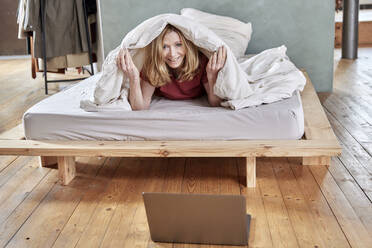 Happy woman lying under blanket on bed at home - FMKF07644