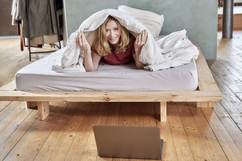 Glückliche Frau liegt unter einer Decke auf dem Bett zu Hause - FMKF07644