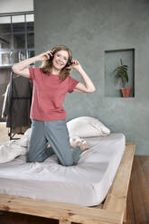 Lächelnde Frau mit drahtlosen Kopfhörern kniend auf dem Bett zu Hause - FMKF07647