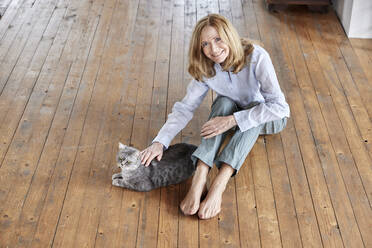 Smiling woman with cat sitting on floor at home - FMKF07650
