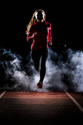 Young woman jogging on running track amidst fog in dark at night - STSF03197
