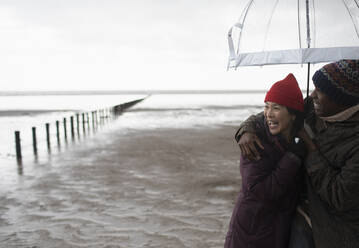 Glückliches Paar, das sich unter einem Sonnenschirm am nassen Winterstrand umarmt - CAIF32342
