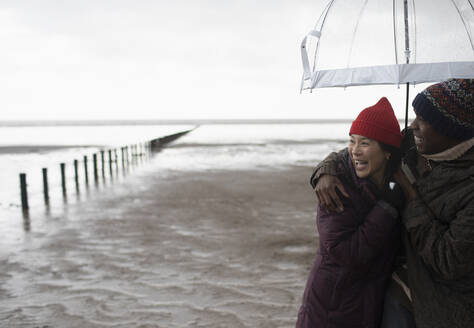 Happy couple hugging under umbrella on wet winter beach - CAIF32342