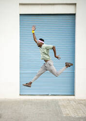 Playful young man jumping mid-air with hand raised on footpath - RCPF01449