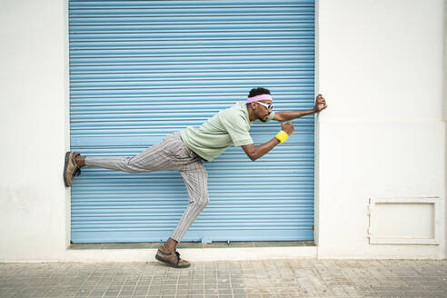 Playful young man standing on one leg in front of blue shutter - RCPF01452