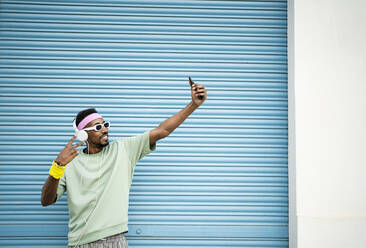 Happy man taking selfie with smart phone and listening music through headphones in front of blue shutter - RCPF01457