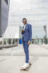 Young businessman using smart phone skateboarding on footpath - IFRF01607