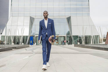 Young businessman holding skateboard and laptop walking in front of office building - IFRF01610