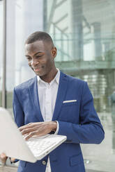 Smiling businessman using laptop in front of modern office building - IFRF01614