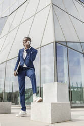 Young businessman holding laptop talking on mobile phone in front of modern building - IFRF01620