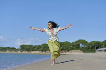 Happy woman running with arms outstretched at beach on sunny day - KIJF04461