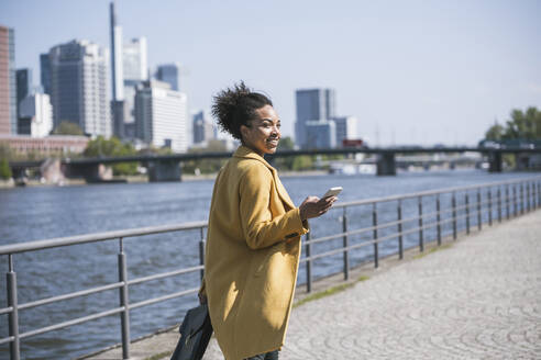 Geschäftsfrau mit Smartphone beim Spaziergang am Mainufer - UUF25962