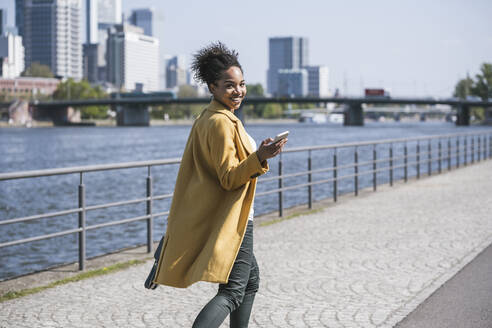 Lächelnde Geschäftsfrau mit Mobiltelefon bei einem Spaziergang am Main an einem sonnigen Tag - UUF25964