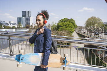 Junge Geschäftsfrau mit Skateboard hört Musik über Kopfhörer an einem sonnigen Tag - UUF25989