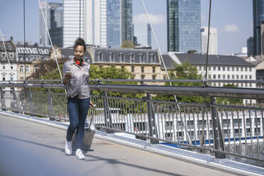 Junge Frau mit Smartphone, die mit Gepäck auf einer Brücke geht - UUF25996