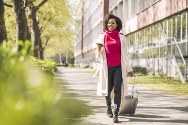 Businesswoman with wheeled luggage and smart phone on footpath - UUF26003