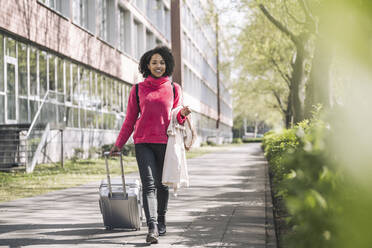 Young businesswoman walking with wheeled luggage on footpath - UUF26005