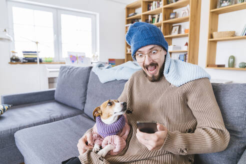 Shocked man with wooly hat sitting on couch holding dog checking smartphone - KMKF01810