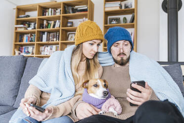 Paar mit Hund sitzt auf der Couch, eingewickelt in eine Decke, und schaut auf sein Smartphone - KMKF01812