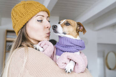 Woman wearing wooly hat at home kissng dog with scarf - KMKF01814