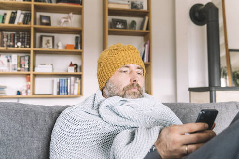 In eine Decke eingewickelter Mann mit Wollmütze sitzt auf der Couch und überprüft sein Smartphone - KMKF01842