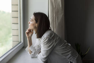 Mature woman with hand on chin looking through window at home - LLUF00502