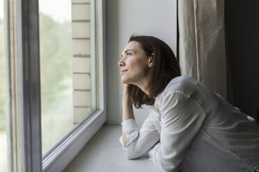 Smiling mature woman looking through window at home - LLUF00503