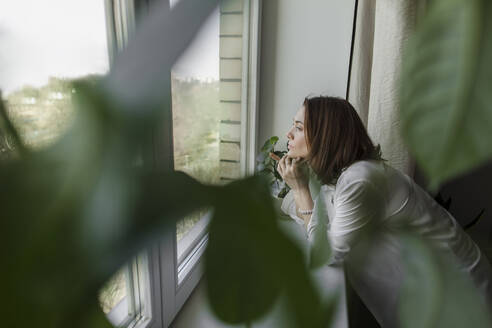 Nachdenkliche Frau mit Hand am Kinn, die durch das Fenster ihres Hauses schaut - LLUF00505