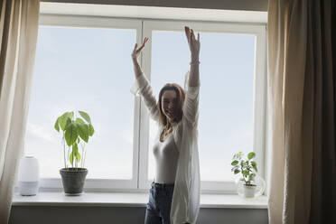 Happy mature woman with arms raised standing in front of window - LLUF00509