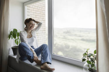 Happy mature woman with hand in hair sitting on window sill at home - LLUF00511