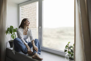 Happy mature woman sitting on window sill at home - LLUF00512