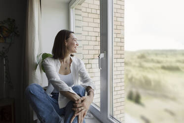 Lächelnde reife Frau, die zu Hause auf der Fensterbank sitzt - LLUF00513